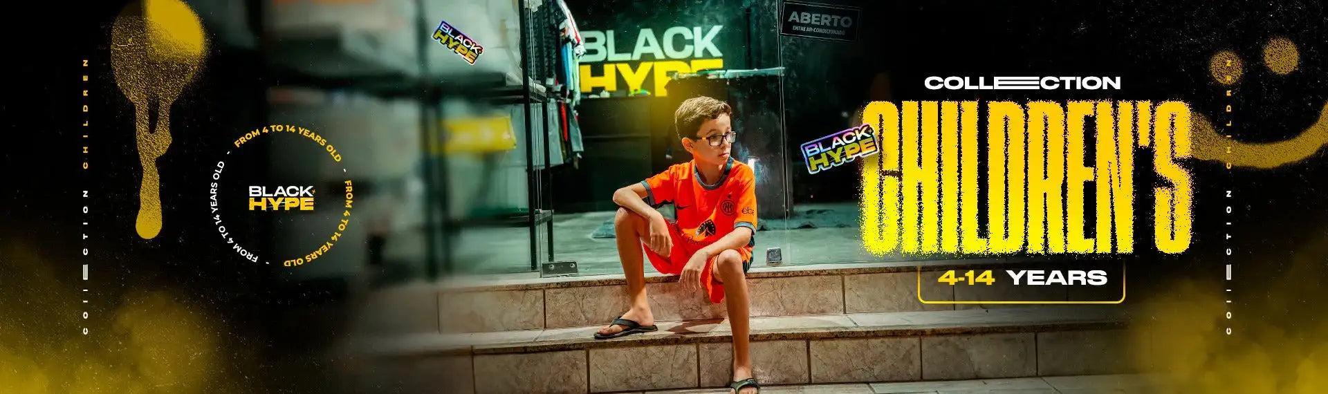A young boy in an orange short-sleeved shirt and matching shorts sits on stone steps, wearing black sandals.